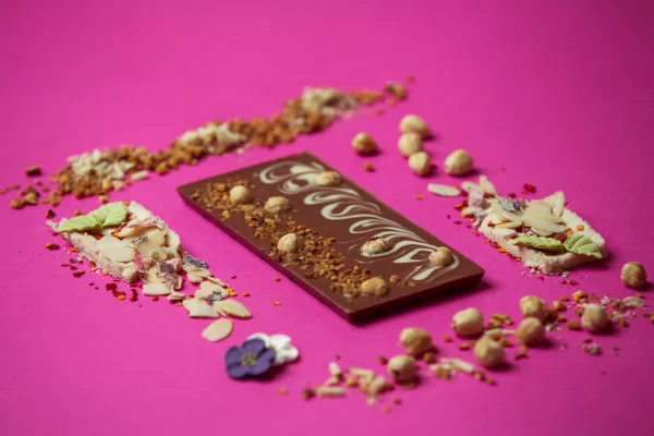 Hazelnut Duet Milk Chocolate bar displayed on a pink background with individual ingredients surrounding.