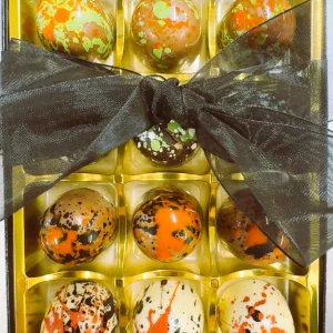 Gold gift box with twelve oval chocolates featuring green, orange, and black splatter patterns, tied with a black organza ribbon.