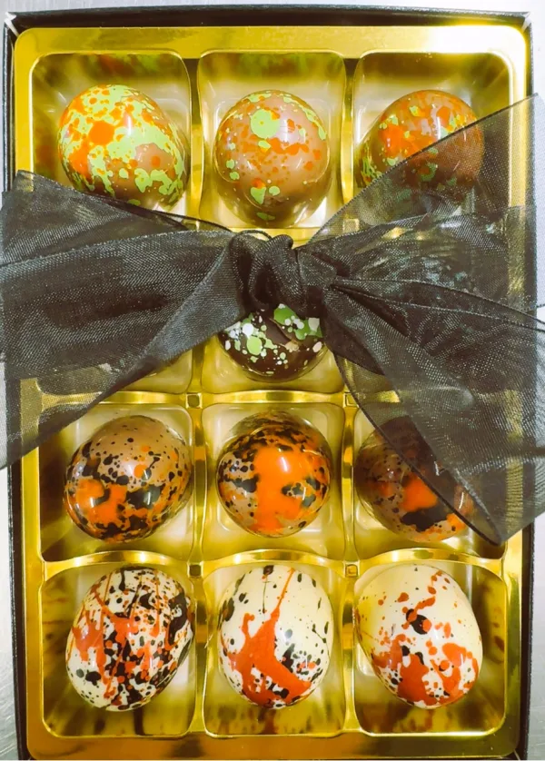 Gold gift box with twelve oval chocolates featuring green, orange, and black splatter patterns, tied with a black organza ribbon.