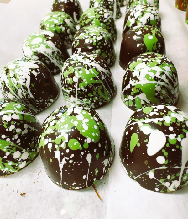 Rows of dome-shaped chocolates with a dark brown coating, decorated with splashes of bright green and white. The chocolates have a shiny finish and are arranged on a white background.