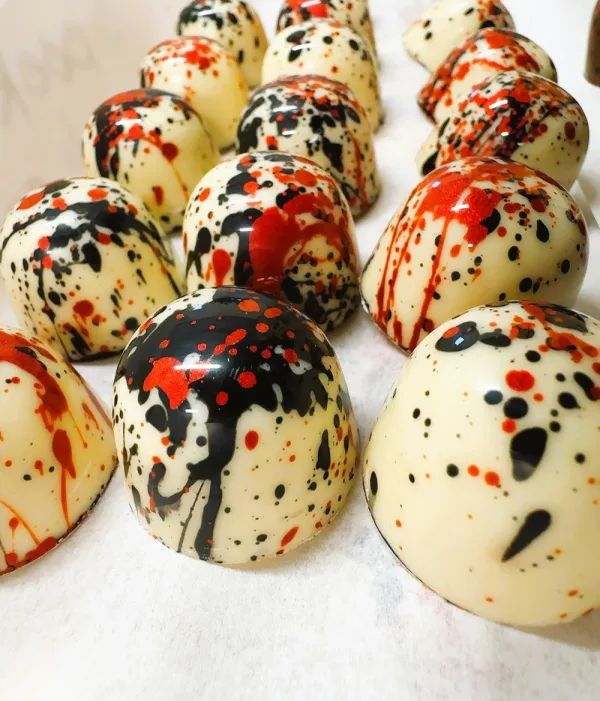 Rows of dome-shaped white chocolates decorated with glossy black and red splatter patterns on a white surface.